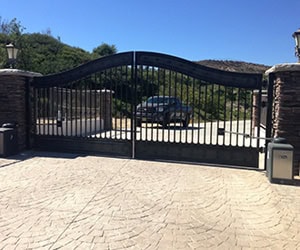 Elegant black wrought iron gate at a residential entrance, showcasing automated opening mechanism, surrounded by stone pillars and a landscaped driveway, reflecting Prime Secured Garage Door & Gate's installation services.