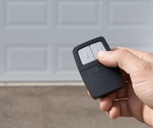 Hand holding a garage door remote control in front of a closed garage door, illustrating remote programming and operation services offered by Prime Secured Garage Door & Gate.
